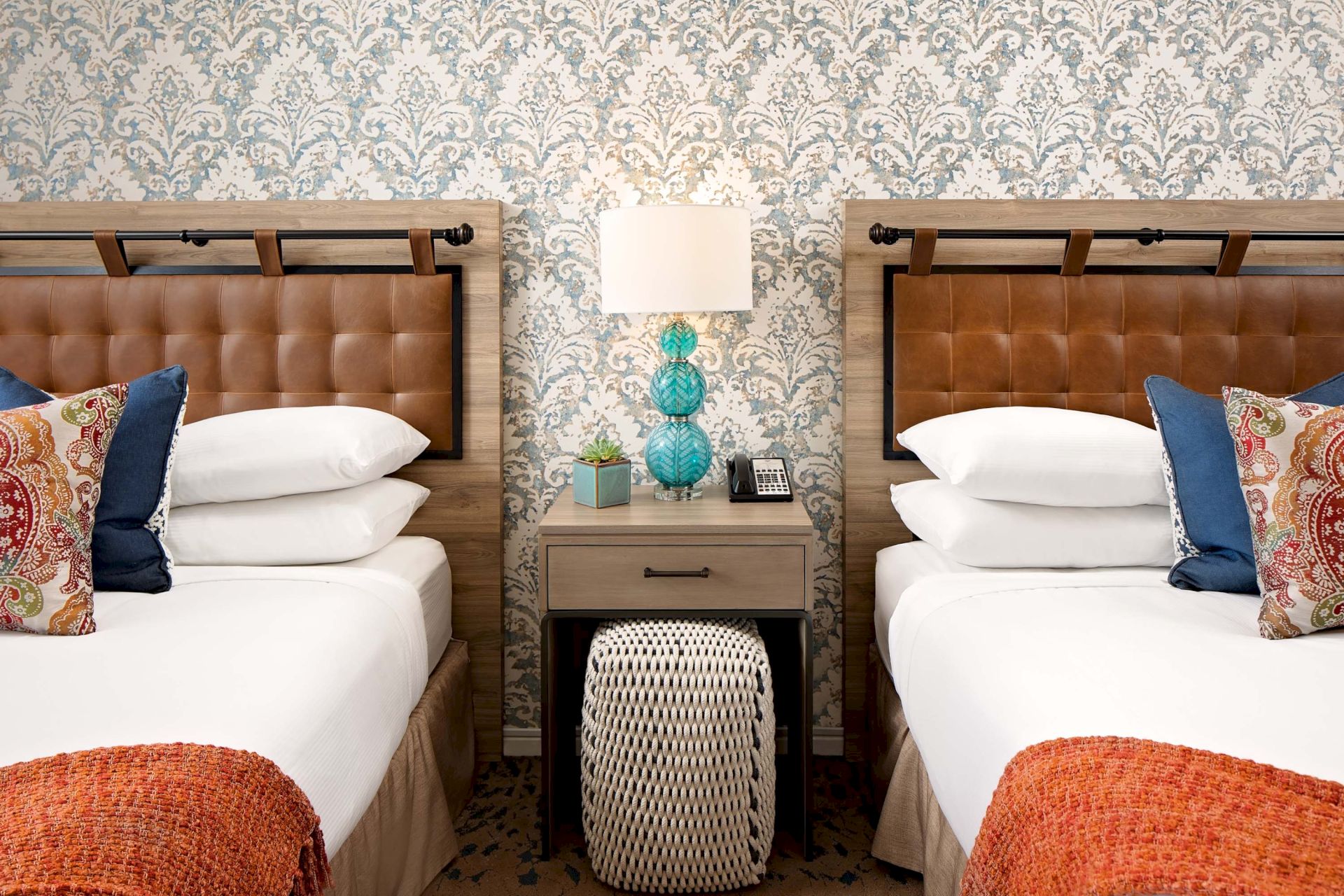 A stylish hotel room shows two twin beds with wooden headboards, patterned wallpaper, and bedside decor with turquoise lamps.