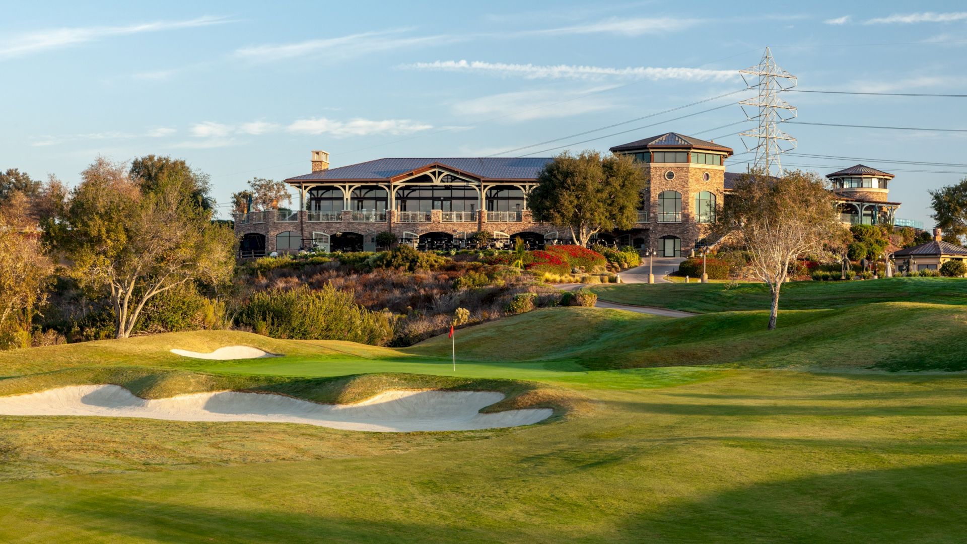 A scenic golf course with a well-manicured green, sand bunkers, and a clubhouse atop a gentle hill, framed by trees under a blue sky.