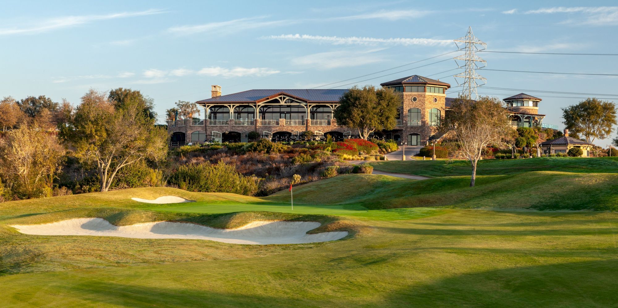 A scenic golf course with a well-manicured green, sand bunkers, and a clubhouse atop a gentle hill, framed by trees under a blue sky.