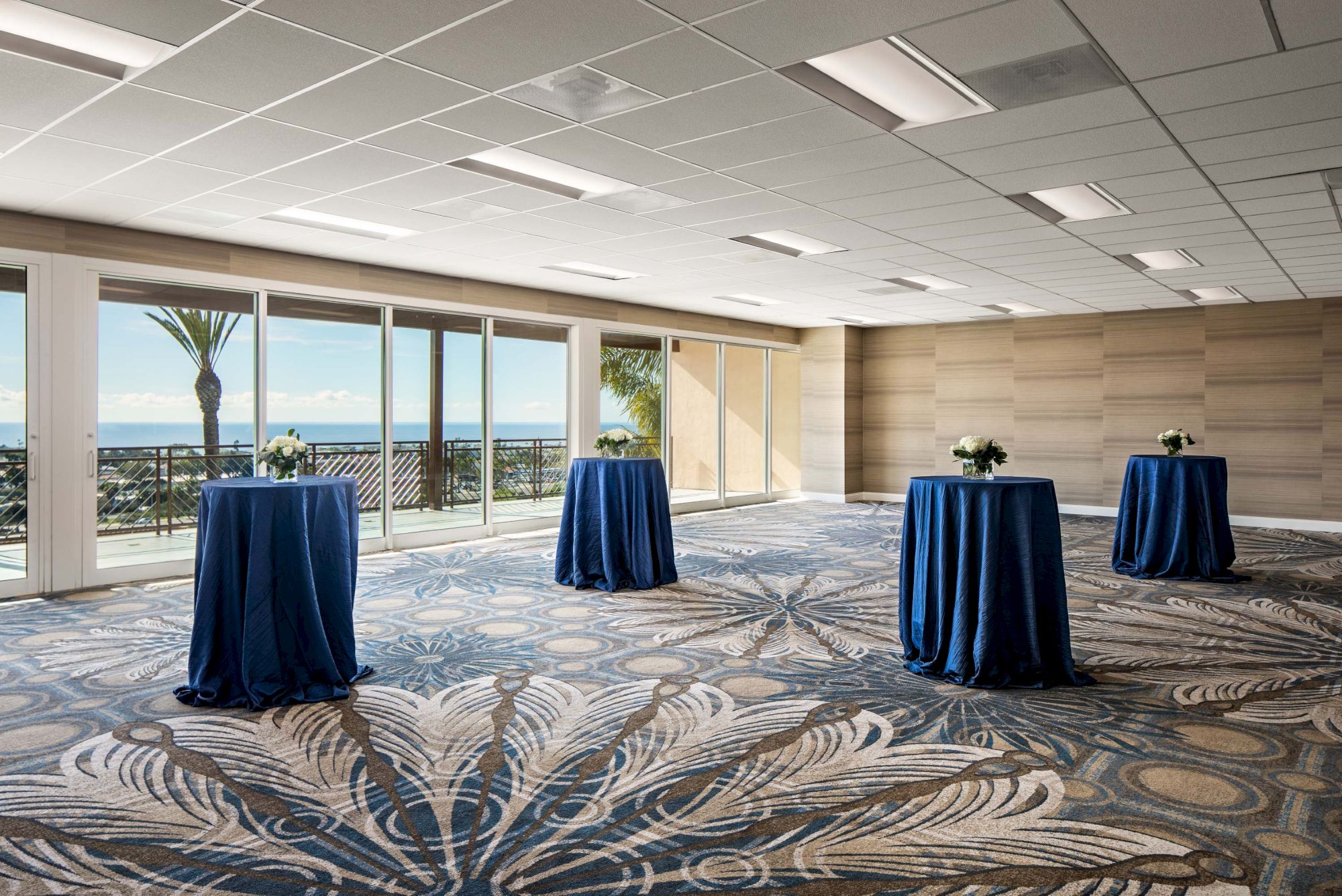 A bright, modern event room with large windows, blue-draped high-top tables, carpeted swirling patterns, and a view of the ocean.