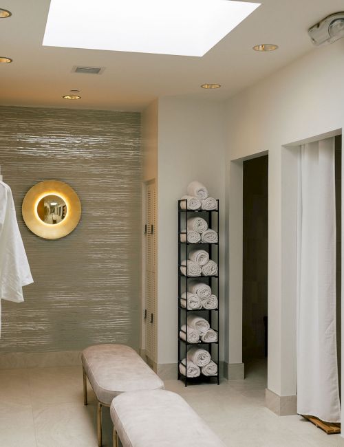 Lockers on left, white robes hanging, circular mirror on tiled wall, benches in center, shelves with towels on right&mdash;spa dressing room.
