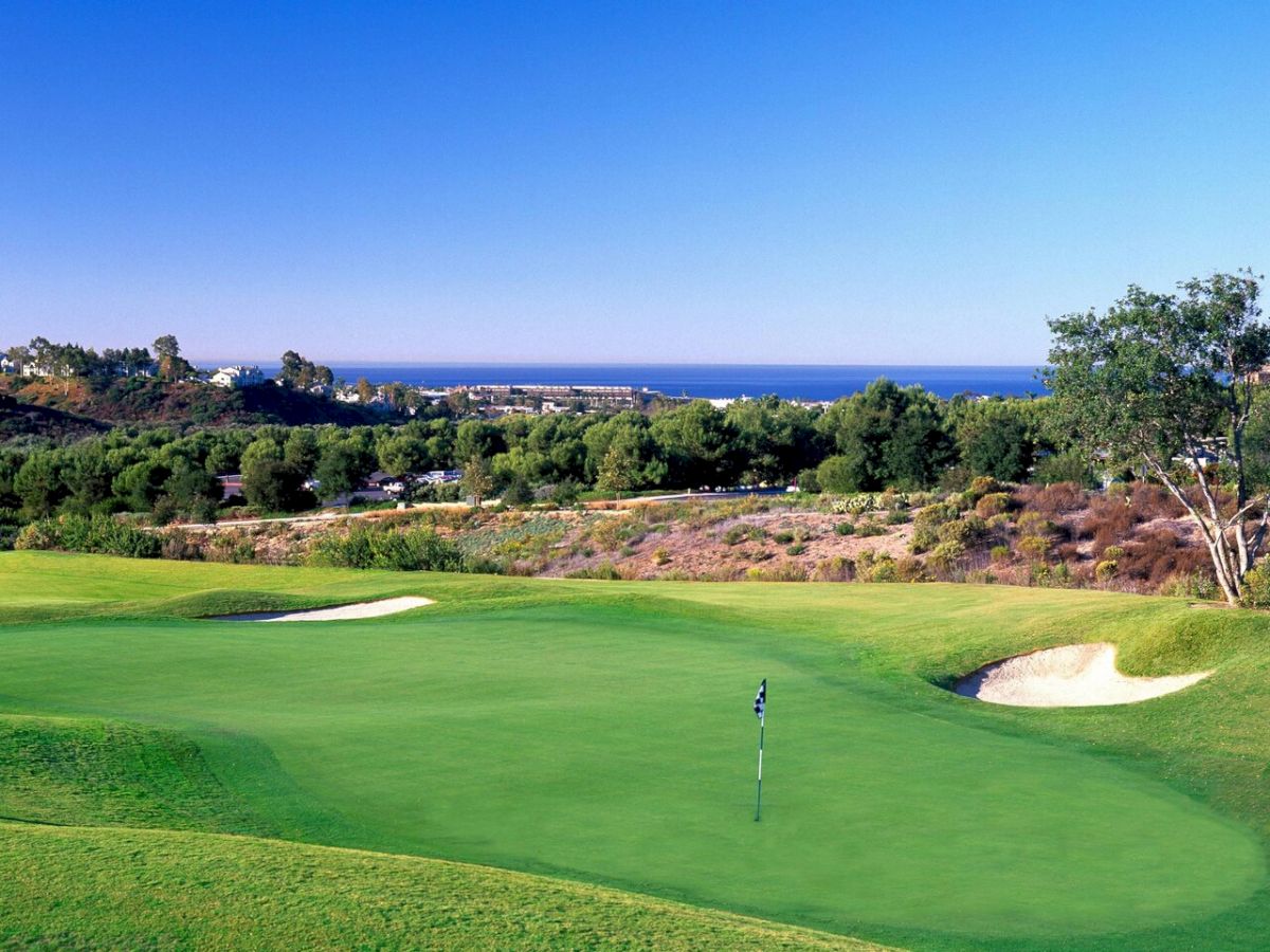 A lush golf course with a green, flagstick, sand bunkers, and rolling hills under a clear blue sky, ocean visible in the distance.