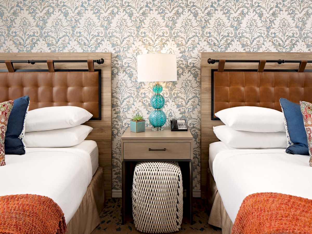 A guest room at The Cassara Carlsbad with two queen beds, tan tufted leather headboards, patterned wallpaper and a turquoise glass bedside lamp.