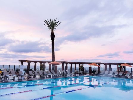 A resort pool with clear blue water, poolside loungers and umbrellas, a tall palm tree, and a pier-like wooden structure against a pastel sunset.
