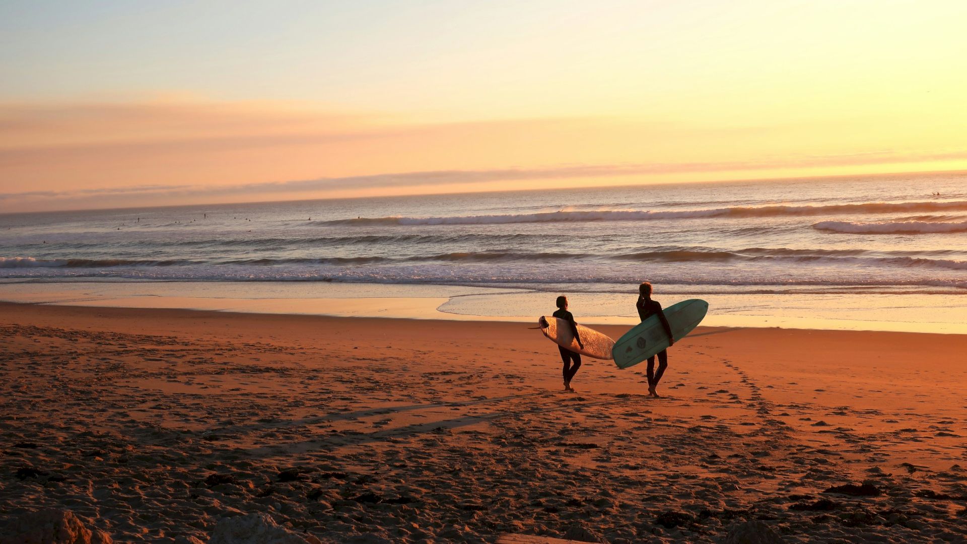 Surfers walk along a sandy beach at sunset, carrying surfboards as waves roll in under a glowing sky.