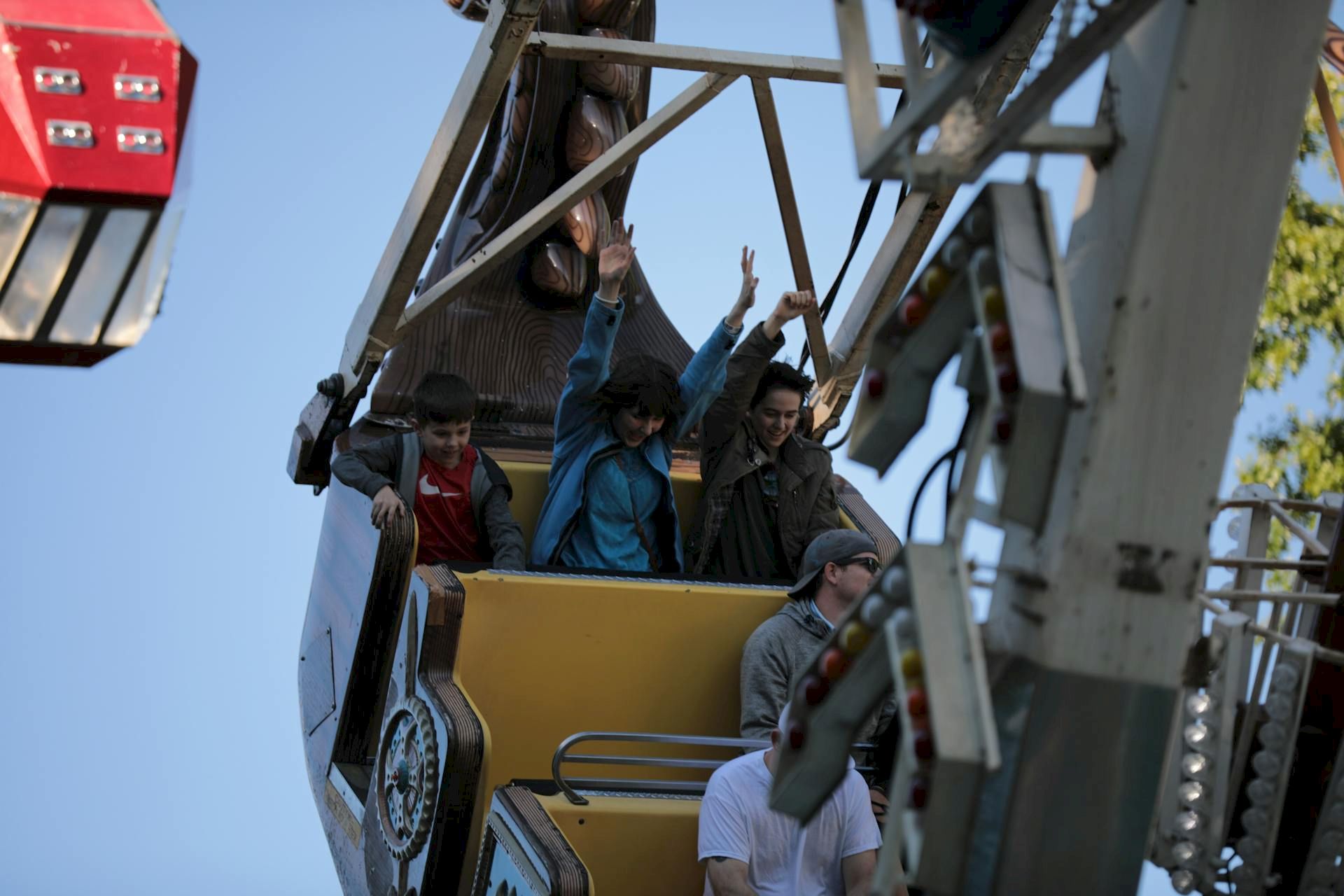 People ride a tilted amusement park ride, looking excited as they lift their arms; a safety harness and support frame are visible.