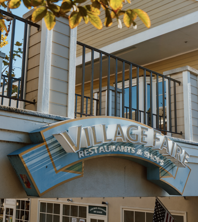A building exterior with an iron balcony railing and a large “Village Fair” sign for restaurants and shops.
