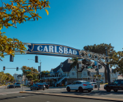 A large “CARLSBAD” street sign spans the road in sunny weather, with trees, traffic lights, and cars nearby.