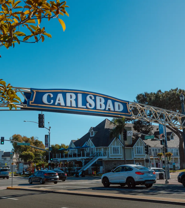 A large “CARLSBAD” street sign spans the road in sunny weather, with trees, traffic lights, and cars nearby.