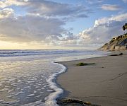 A sandy beach with gentle waves, palm trees on cliffs, and two people walking under a cloudy, sunset sky.