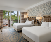 A bright hotel room with two neatly made twin beds, patterned wallpaper, warm lamps, and curtains opening to a palm-filled view.