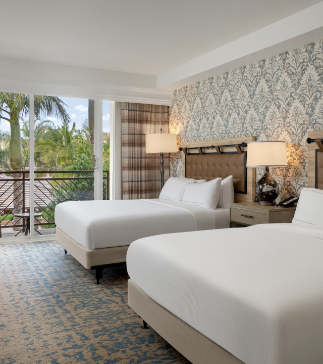 A bright hotel room with two neatly made twin beds, patterned wallpaper, warm lamps, and curtains opening to a palm-filled view.