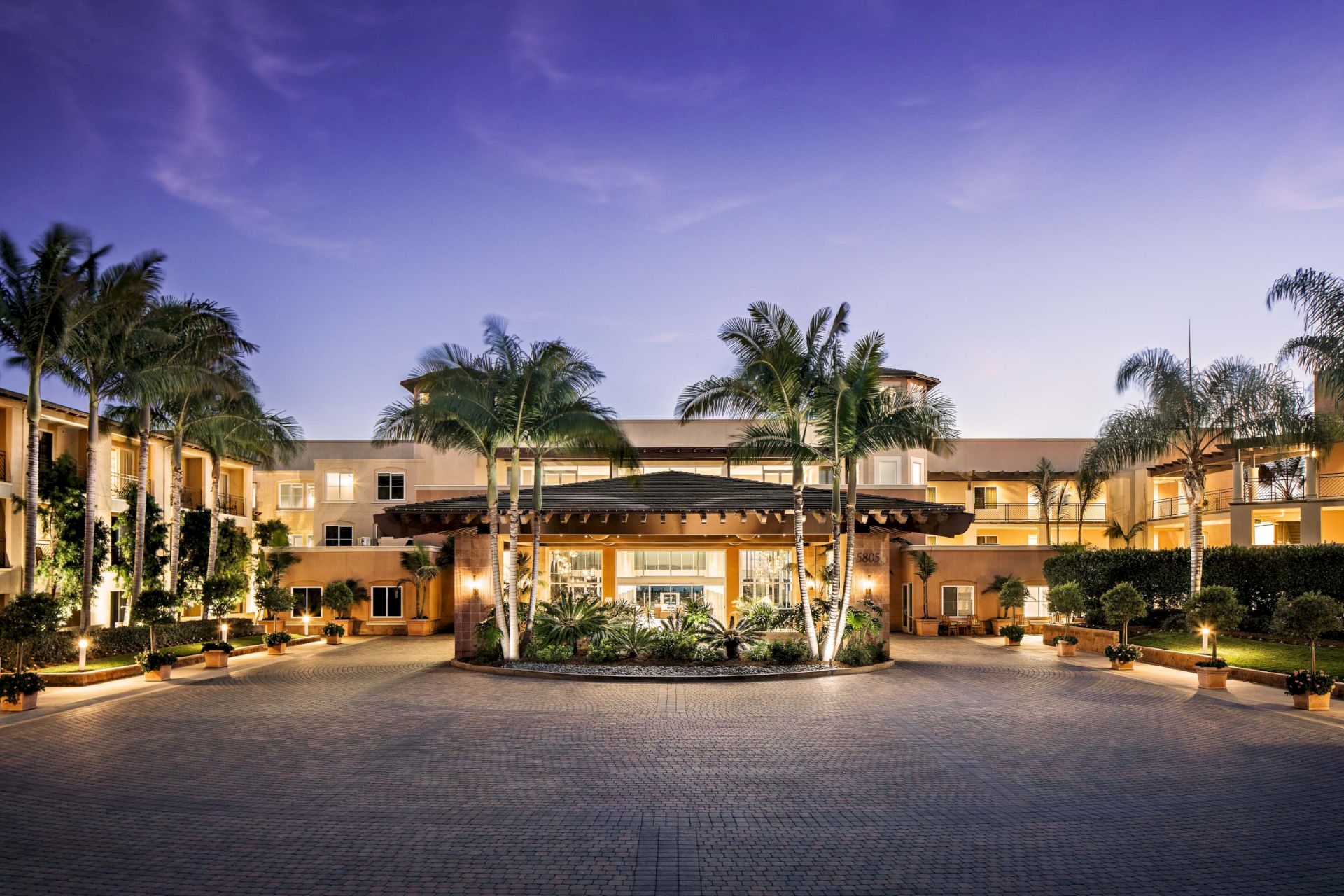 A warm evening view of a hotel courtyard with palm trees, landscaped gardens, and lit buildings under a purple sky.