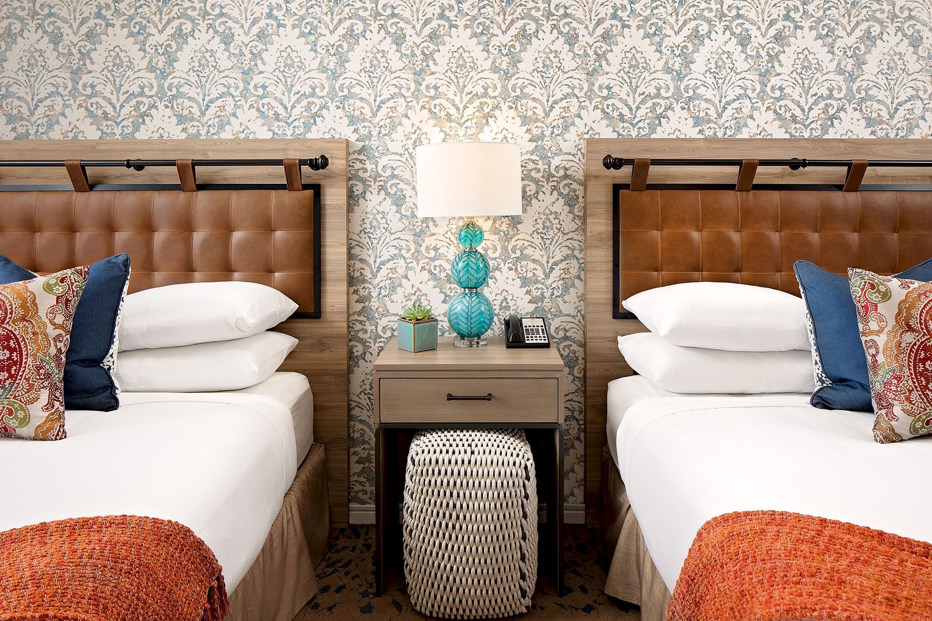 Two twin beds with tufted headboards, a shared nightstand, a turquoise lamp, patterned wallpaper, and decorative pillows in a cozy hotel room.