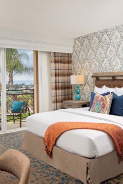 A bright hotel room with a king bed, patterned wallpaper, teal lamps, and a balcony view of palm trees and the ocean, ending with a neat finish.