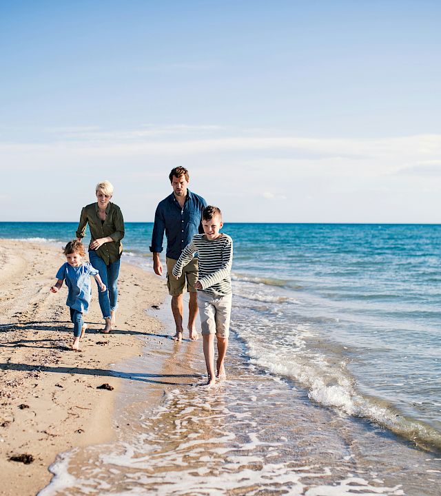 A family of four walks along a sunny beach, waves lapping at their feet, as they enjoy a seaside stroll together.