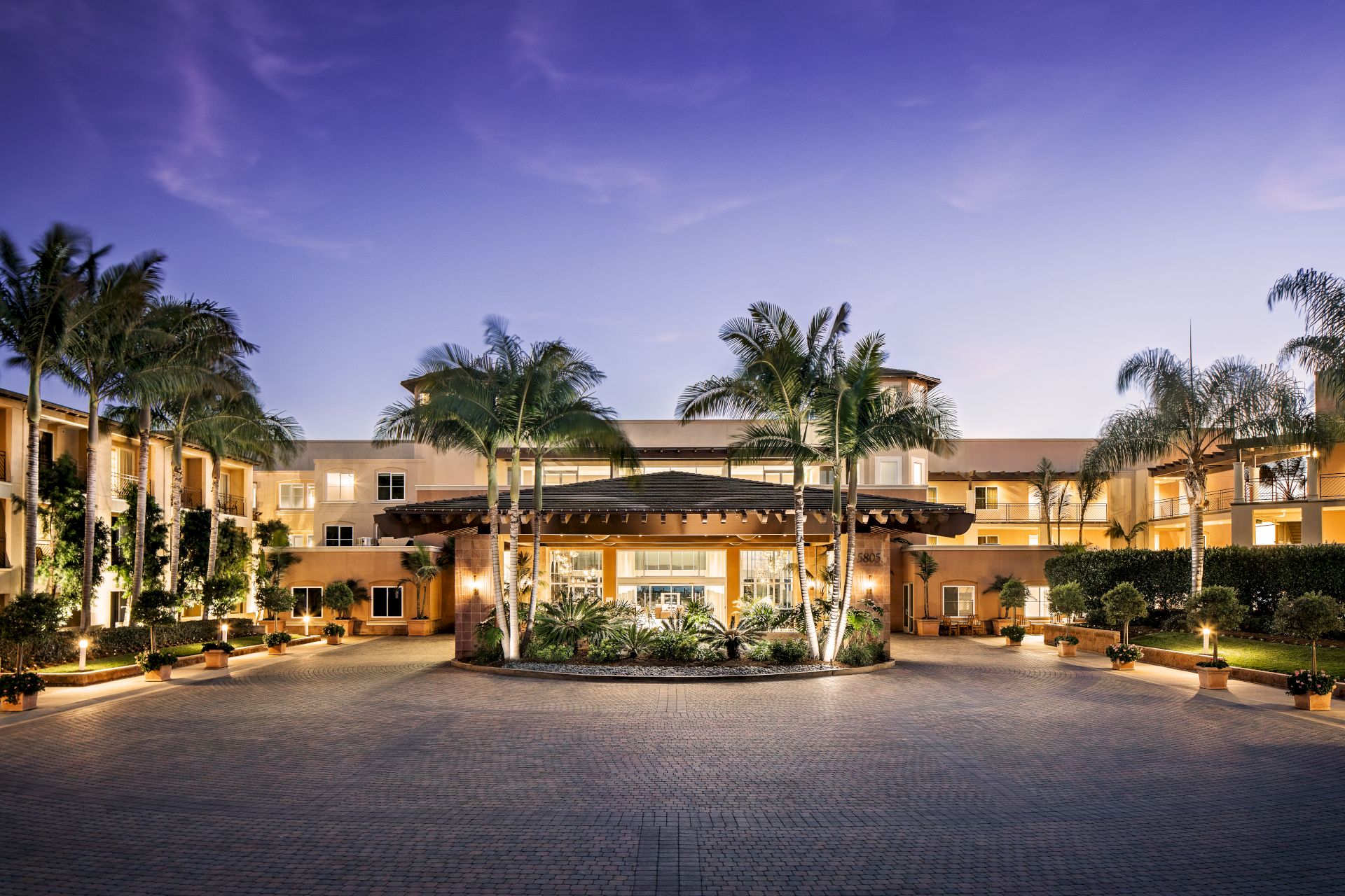 A lit resort hotel at dusk with palm trees and a landscaped courtyard under a purple sky.